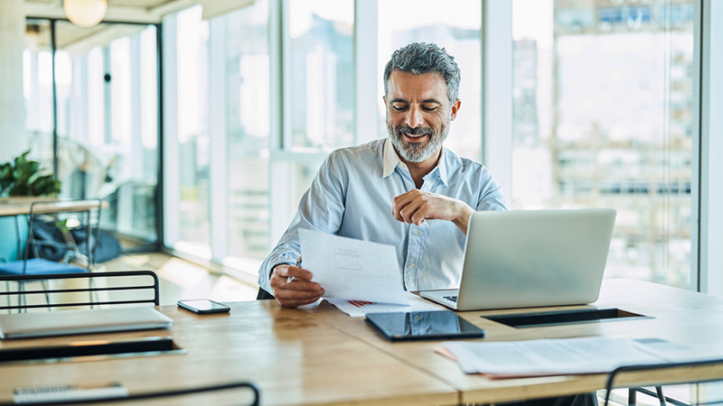 Man looks over a financial sheet while sitting in an office with his laptop open